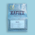 A bag of KAFiEX ROASTERS Brazil whole bean coffee on a solid light blue background. The packaging is light blue with white botanical line art, featuring the brand name 'KAFiEX ROASTERS' in dark purple text and a white label detailing the coffee's origin and roast.