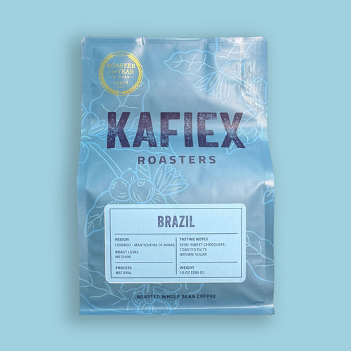 A bag of KAFiEX ROASTERS Brazil whole bean coffee on a solid light blue background. The packaging is light blue with white botanical line art, featuring the brand name 'KAFiEX ROASTERS' in dark purple text and a white label detailing the coffee's origin and roast.