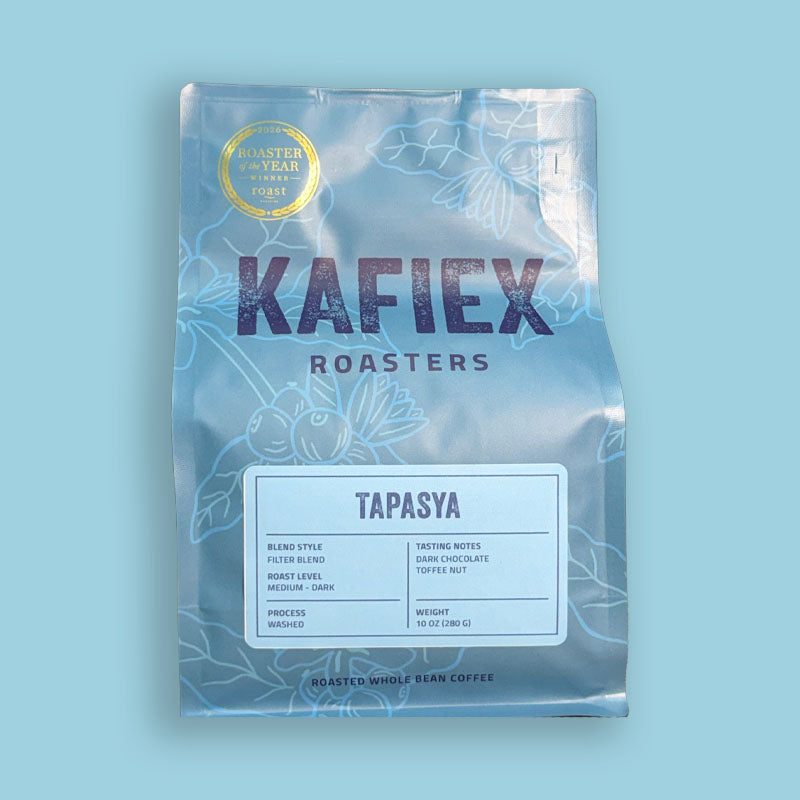 A bag of KAFiEX ROASTERS Tapasya whole bean coffee on a solid light blue background. The packaging is a metallic light blue-gray with white floral line art, featuring the brand name 'KAFiEX ROASTERS' in dark purple and a gold 'Roaster of the Year 2024' seal in the top left corner.