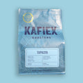 A bag of KAFiEX ROASTERS Tapasya whole bean coffee on a solid light blue background. The packaging is a metallic light blue-gray with white floral line art, featuring the brand name 'KAFiEX ROASTERS' in dark purple and a gold 'Roaster of the Year 2024' seal in the top left corner.